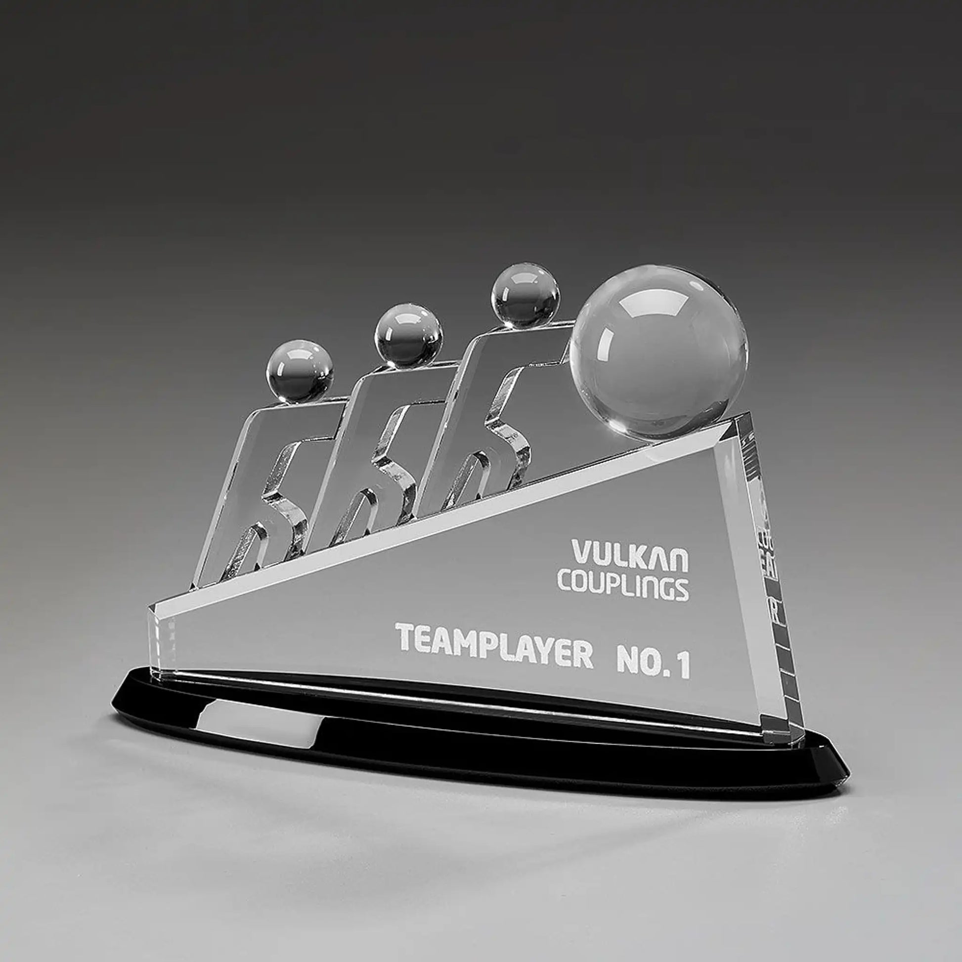 Collaboration Award aus Kristallglas – drei aufstrebende Figuren mit großer Glaskugel auf dreieckigem Glaskörper und schwarzem Sockel, Höhe 145 mm – mit Beispielgravur „Teamplayer No. 1"
