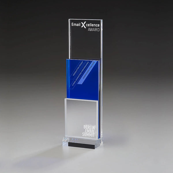 Modern Step Award mit Lasergravur und Digitaldruck: klares Acrylpanel mit Logo und Auszeichnungstitel, blaues Mittelelement mit Kategorietext, mattiertes silberweißes Element mit Veranstaltungsname, klarer Sockel 
