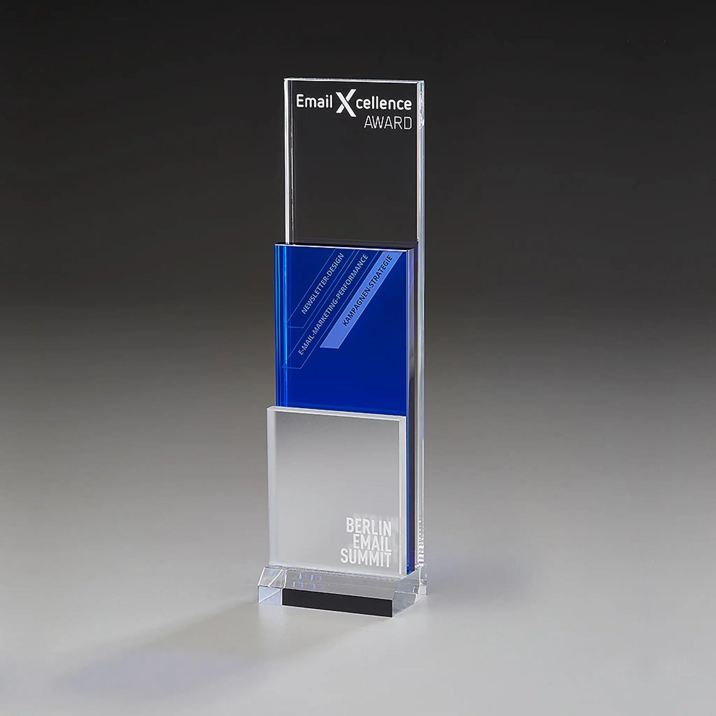 Modern Step Award mit Lasergravur und Digitaldruck: klares Acrylpanel mit Logo und Auszeichnungstitel, blaues Mittelelement mit Kategorietext, mattiertes silberweißes Element mit Veranstaltungsname, klarer Sockel 
